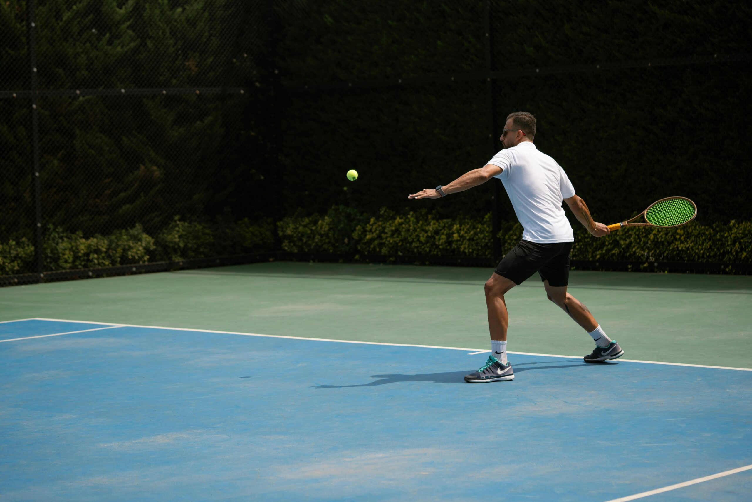 Tennis player hitting a backhand shot on a sunny outdoor court in Iran.